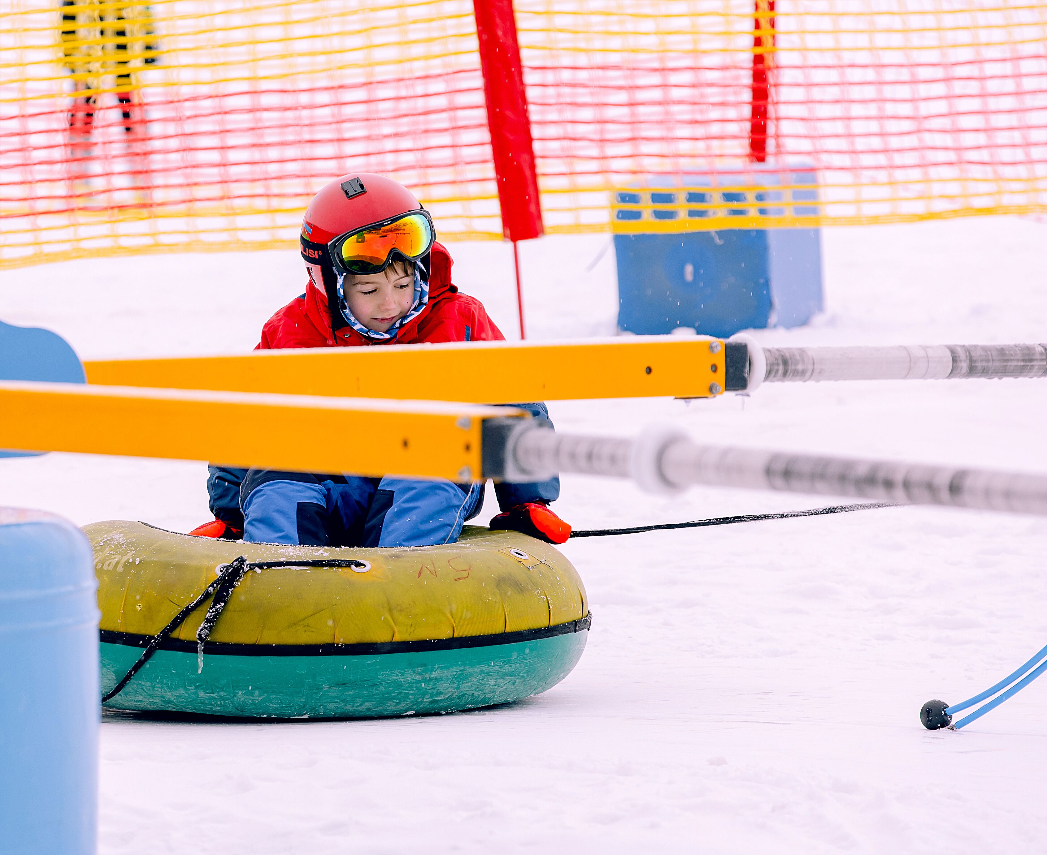 A person in a red jacket and helmet sitting on a yellow inflatable tube in a snowy area with a netted enclosure in the background.
