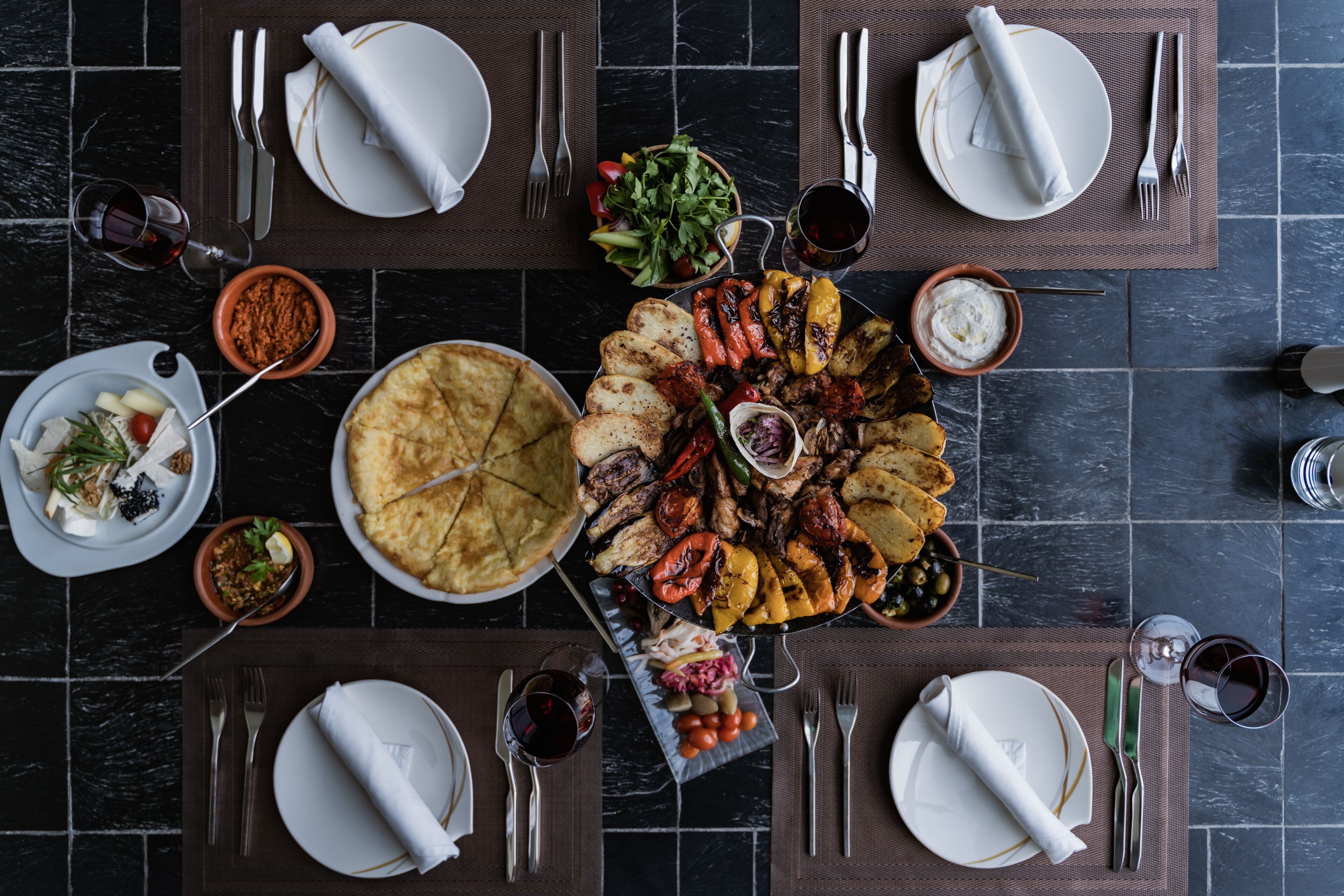 Una mesa lista para una comida con varios platos, incluyendo carnes a la parrilla, verduras y una tarta, sobre un suelo de baldosas oscuras.