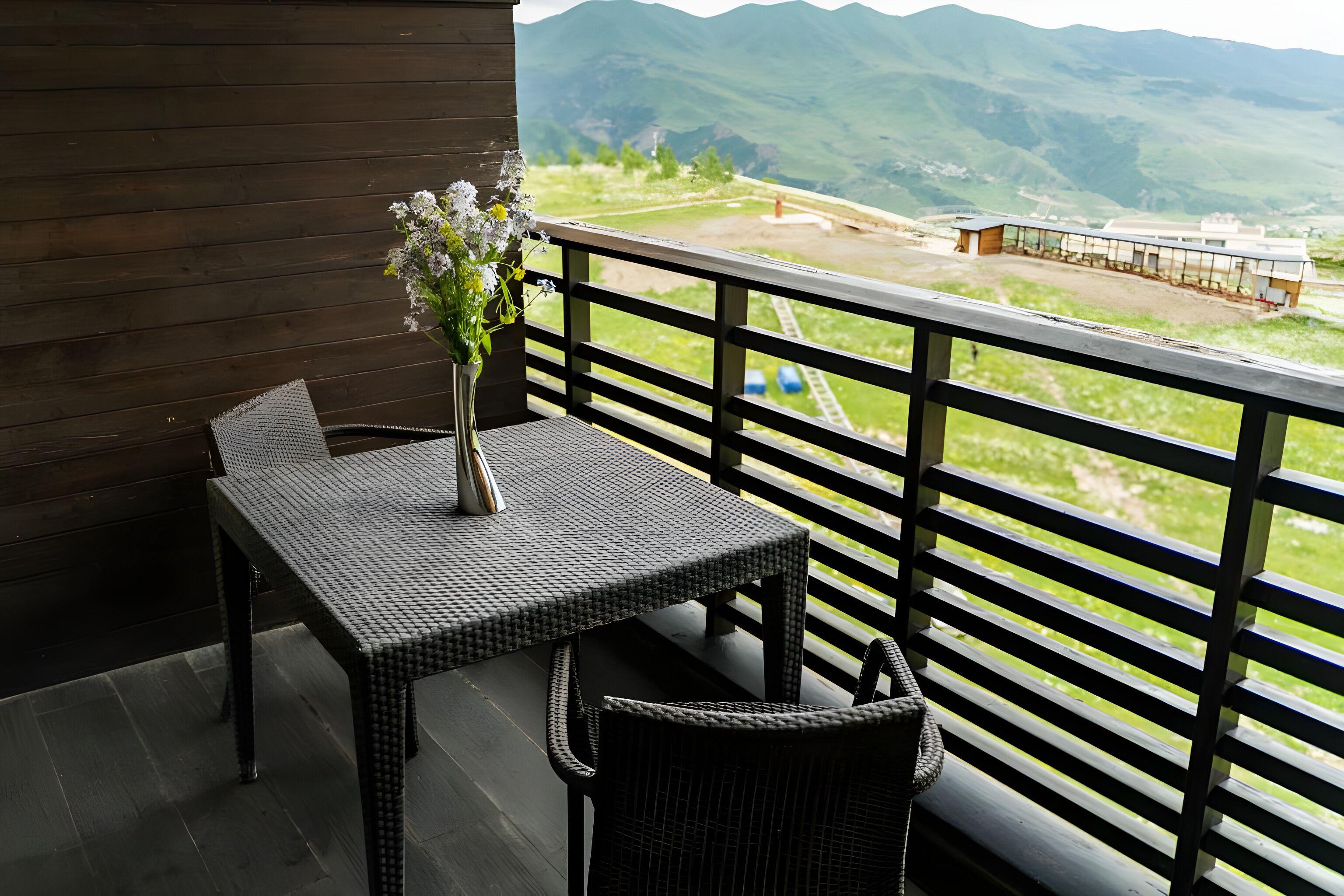 A balcony with a table and chair, a vase with flowers, and a view of a mountainous landscape.