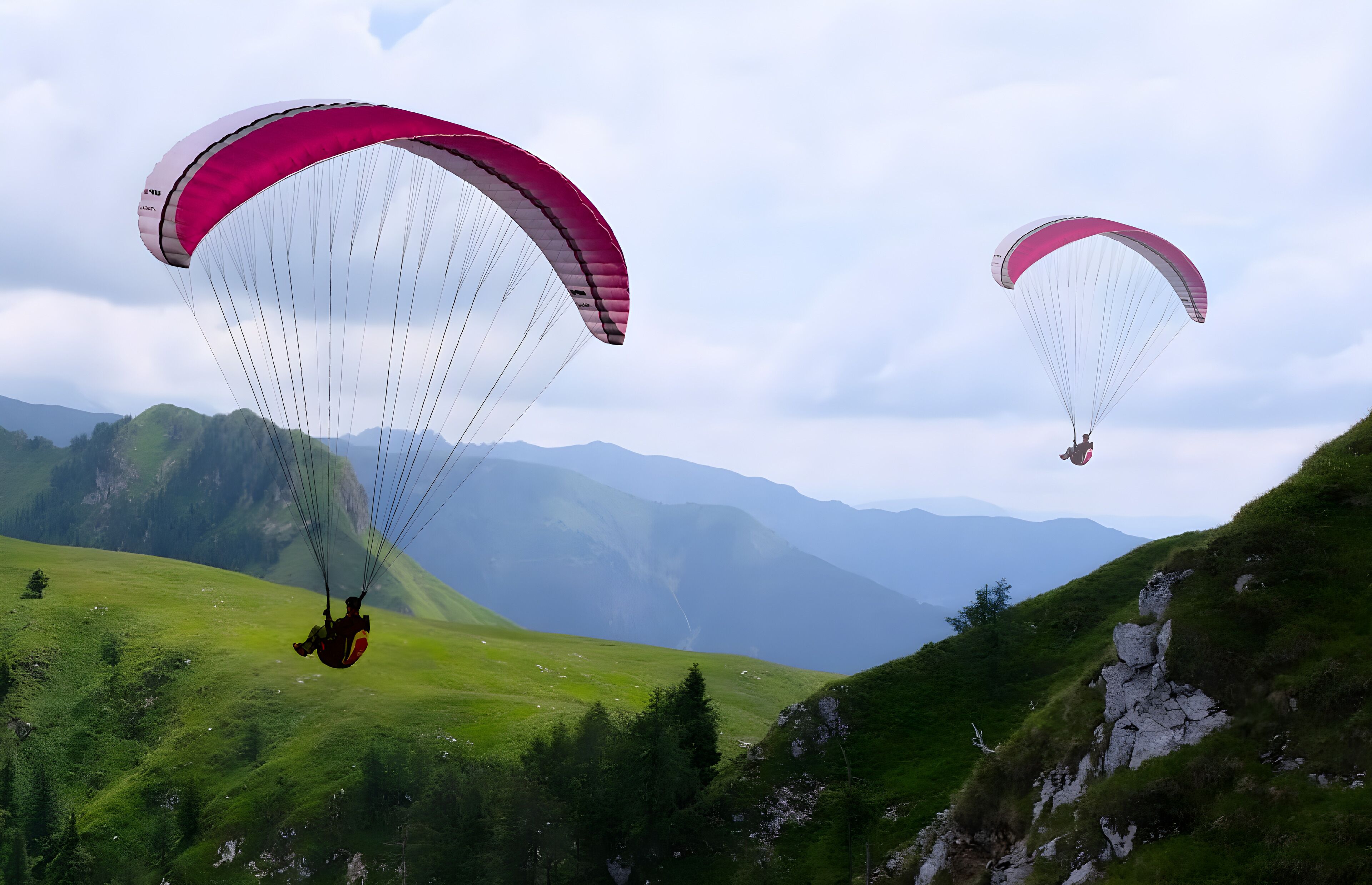 Deux parapentistes évoluent dans un paysage montagneux, avec des collines verdoyantes et des montagnes à l’horizon.
