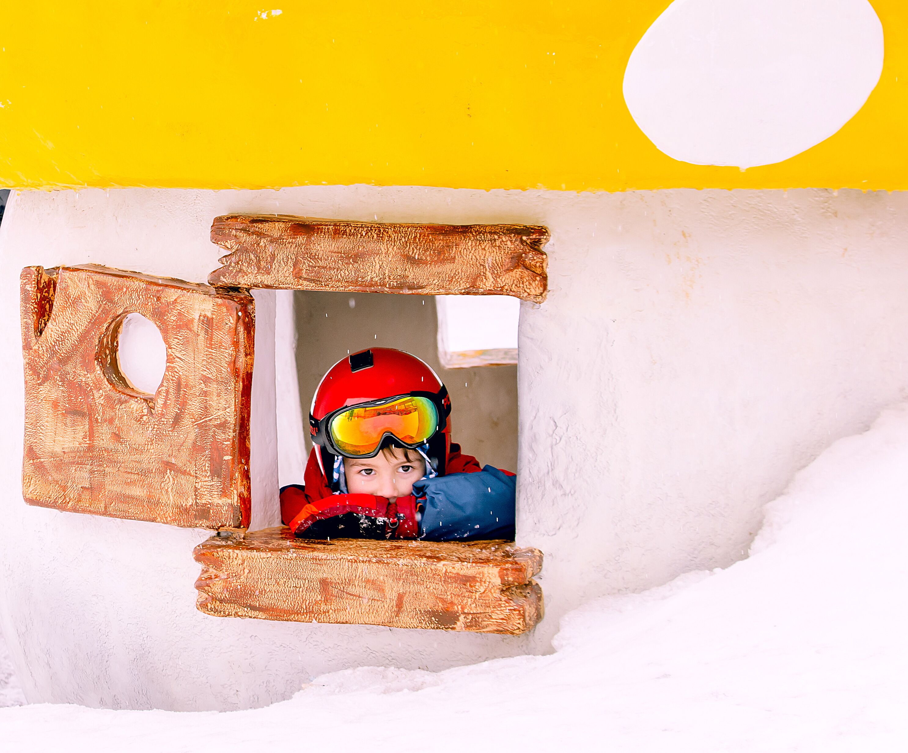 Un niño con casco y gafas de nieve dentro de una cabaña de nieve con techo amarillo y paredes blancas.