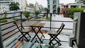 Un balcón con dos sillas de madera y una mesa de vidrio, con vista a un paisaje urbano con edificios modernos y una calle.