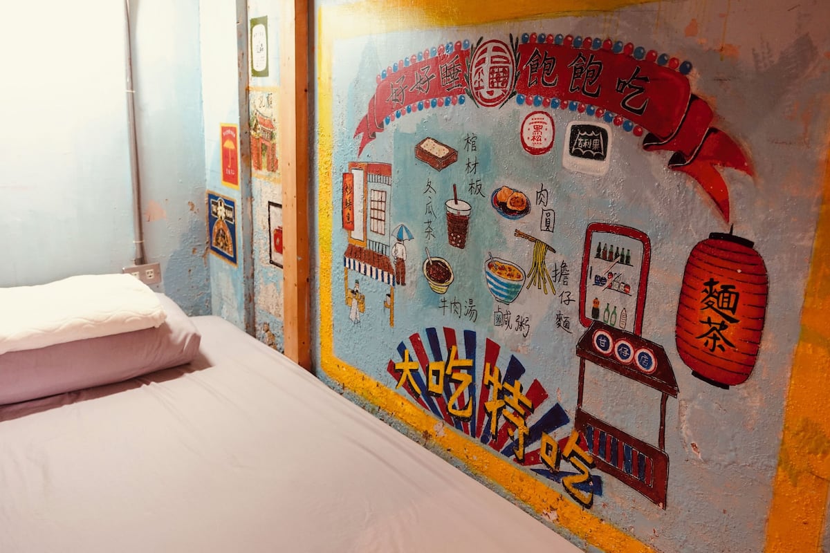 A room with a bed, a wall with colorful artwork depicting food and drinks, and a window with a view of the outside.