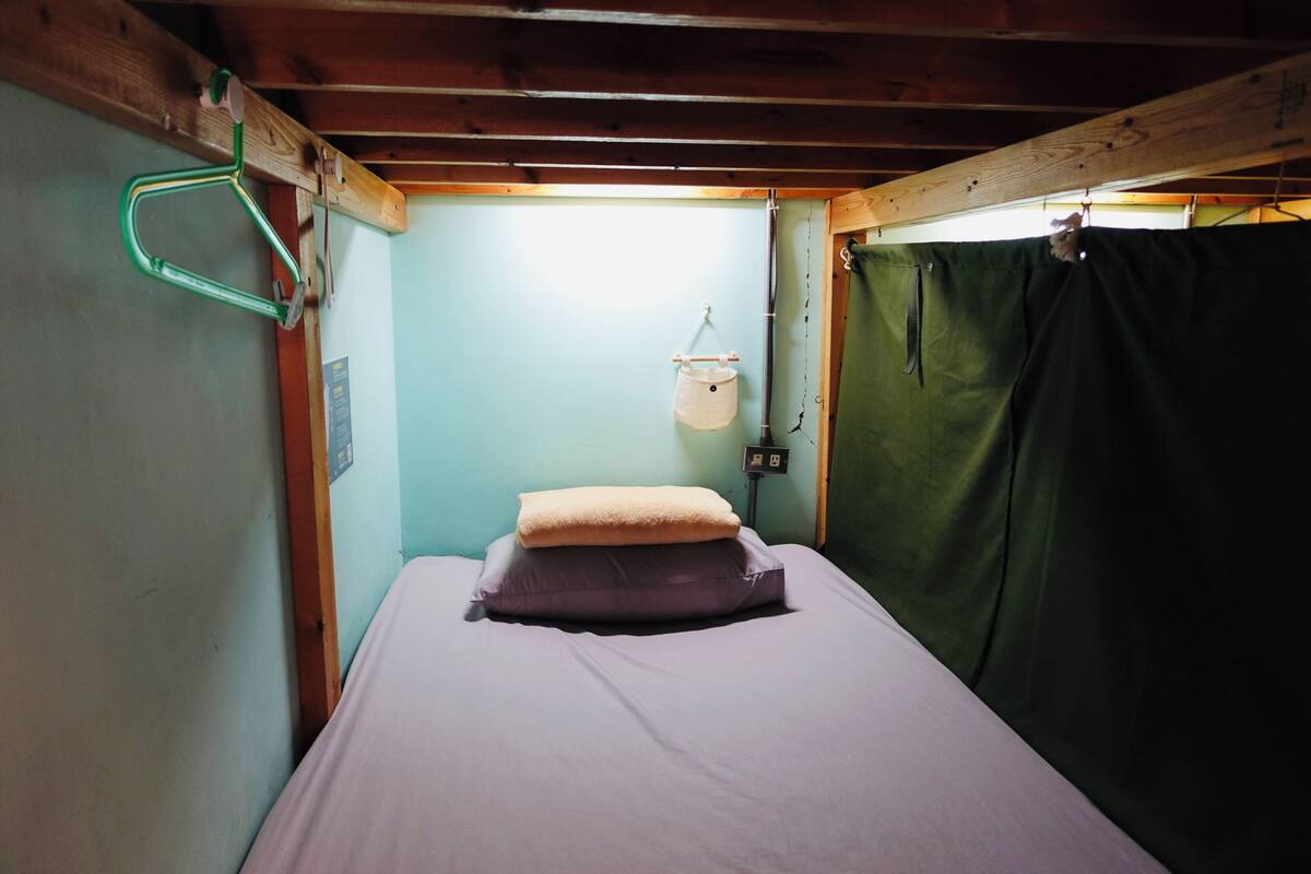A small, narrow room with a single bed, a hanging light fixture, and a green curtain.