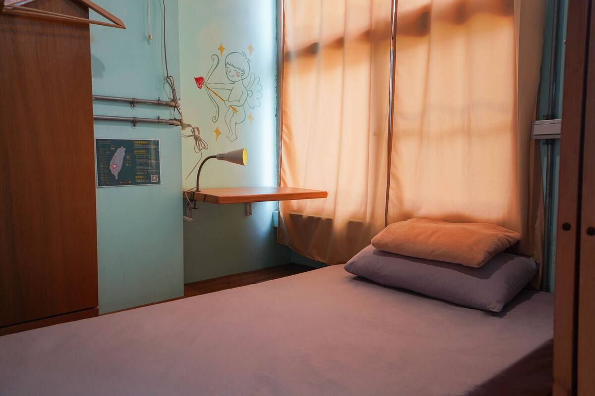 A small, single-bed room with a wooden wardrobe, a map on the wall, and a bedside lamp.