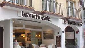 Un hotel con cartel de 'cafetería sucursal', mesas al aire libre con sillas blancas y un balcón con barandilla de metal.