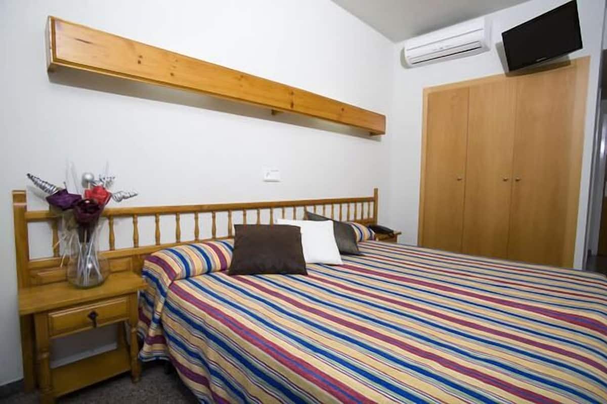 Een slaapkamer met een houten bedframe, een kleurrijk gestreept bedlaken, een nachtkastje met een vaas en een garderobe.
