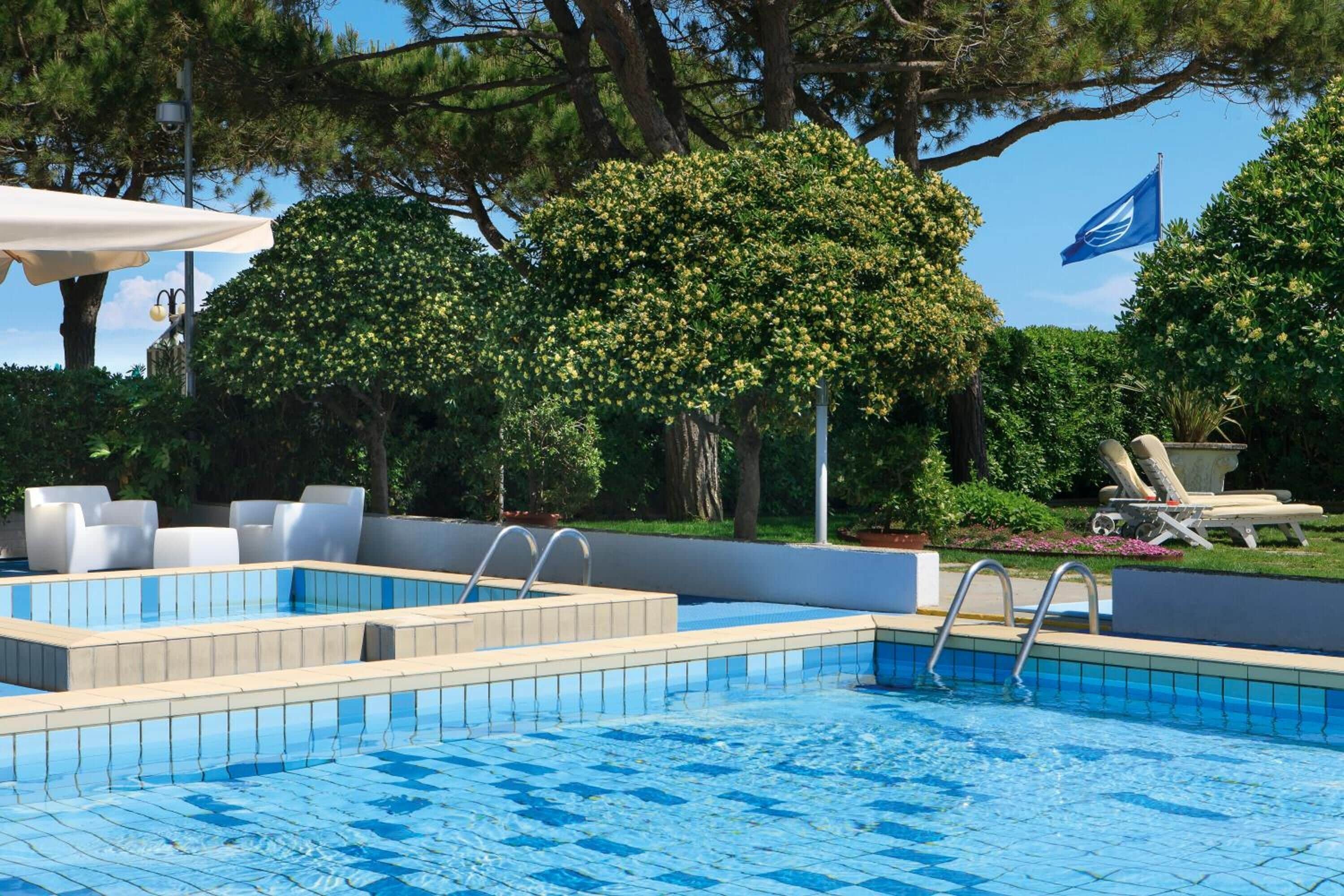 Une piscine dont les bords sont recouverts de carreaux, entourée de chaises longues et d’un grand arbre.