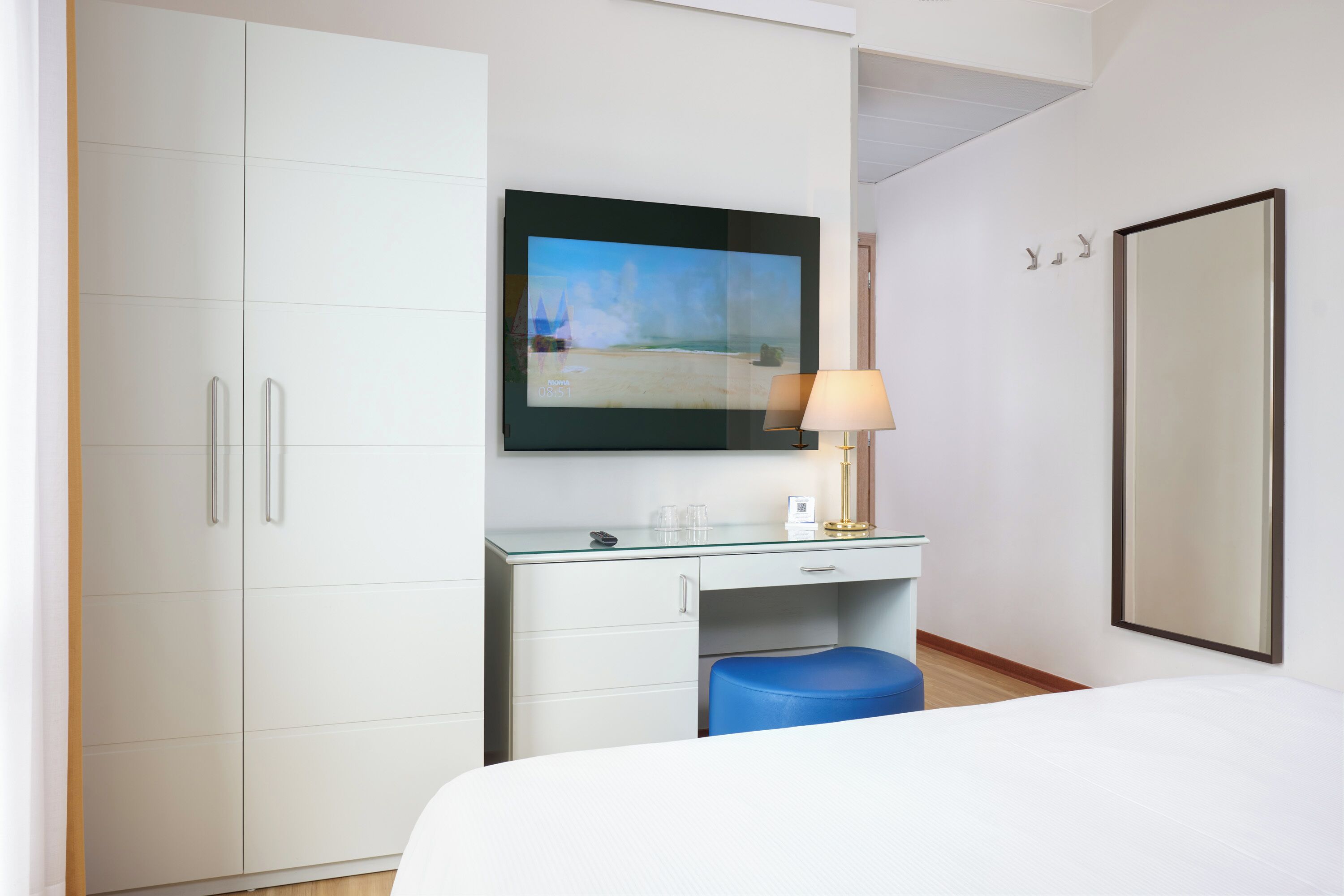 Una camera d'albergo con un letto, una scrivania con una lampada, uno specchio grande e una TV che mostra una spiaggia.