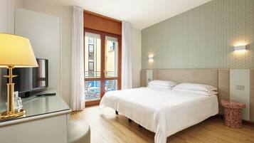 A hotel room with a bed, a desk with a lamp, a television, and a window with curtains.