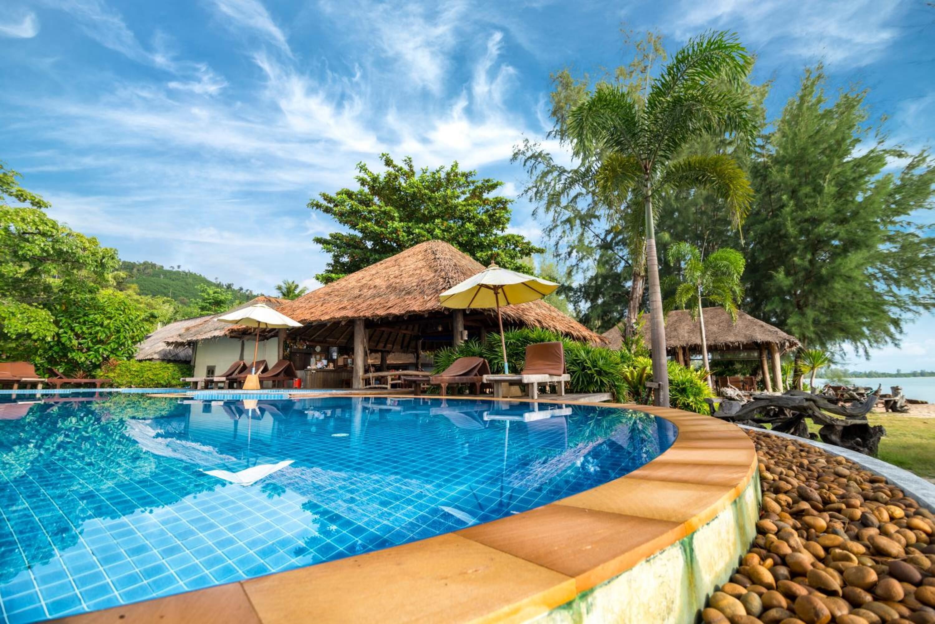 A swimming pool with lounge chairs and umbrellas, surrounded by trees and a thatched-roof building.