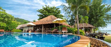 A swimming pool with lounge chairs and umbrellas, surrounded by trees and a thatched-roof building.