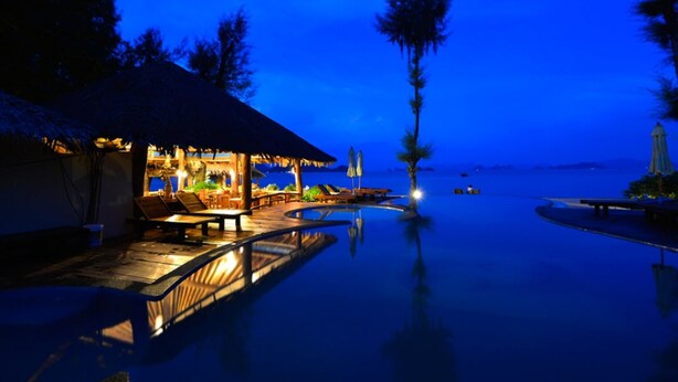 Un resort nocturno con piscina infinita, sillones de descanso y una estructura de techo de paja.