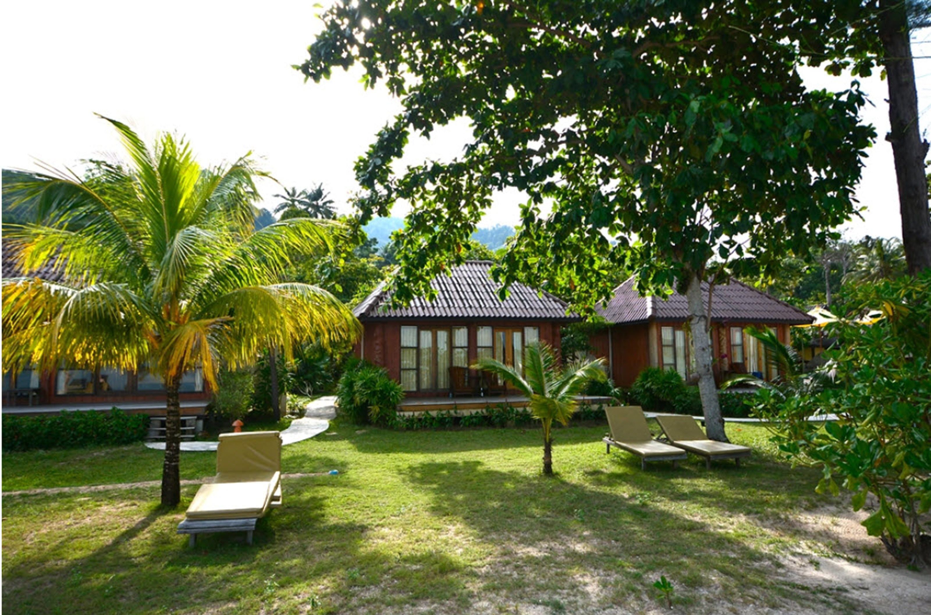 Ein Bungalow mit Ziegeldach, umgeben von üppiger Vegetation und Palmen. Liegestühle und ein kleines Schwimmbecken sind vorhanden.