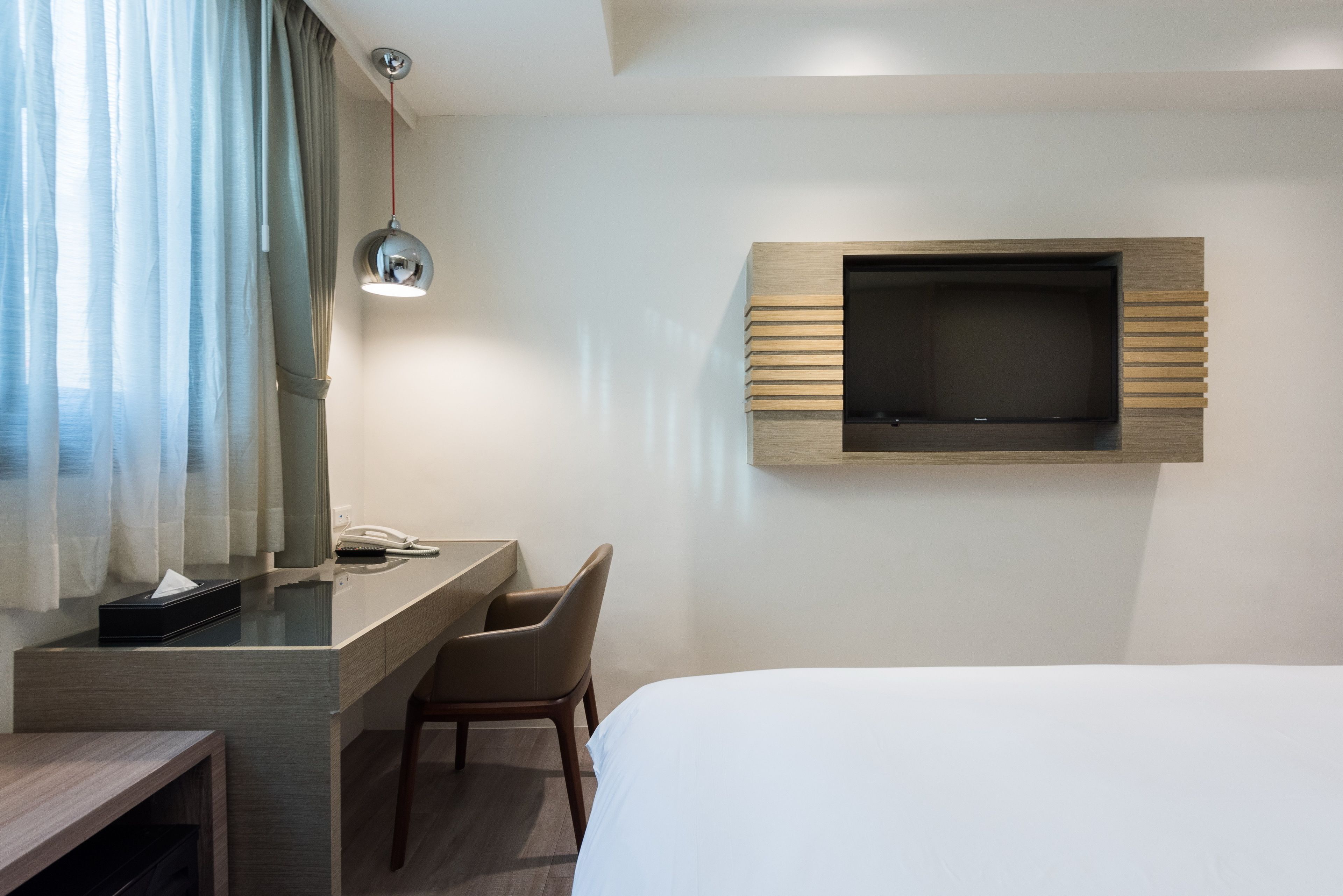 A hotel room with a bed, a desk with a chair, a TV mounted on the wall, and a bedside table with a lamp.