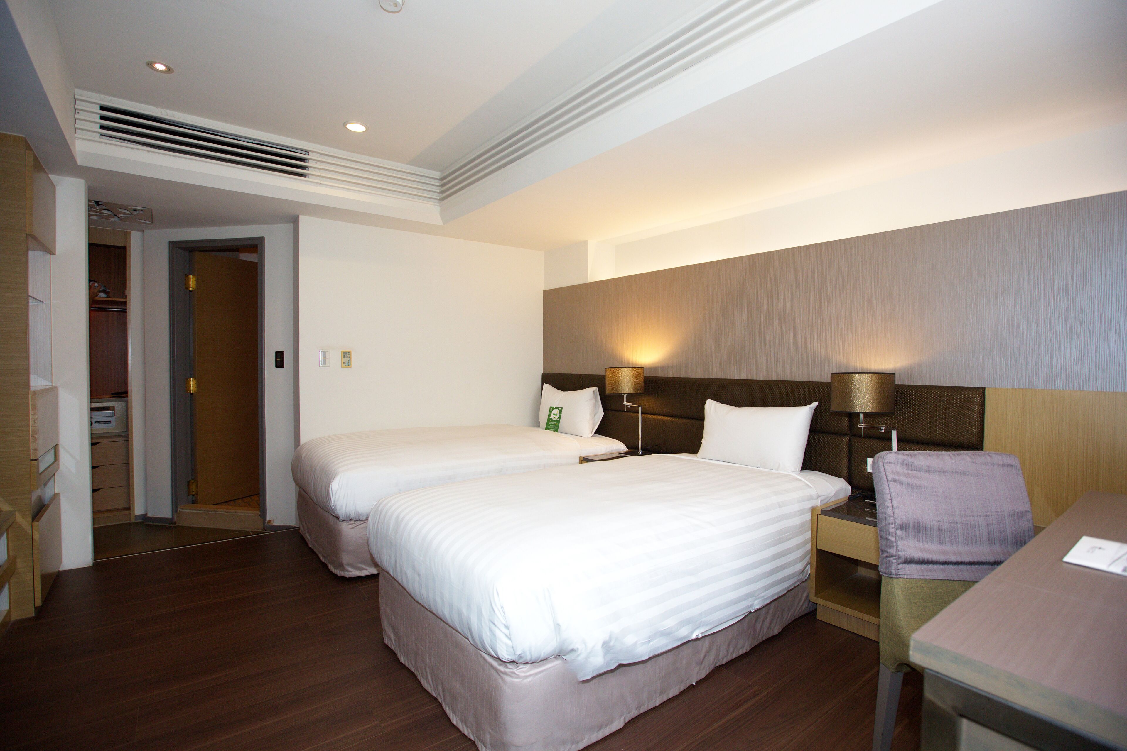 A hotel room with two beds, a desk, and a chair. There is a wardrobe and a door leading to another room.
