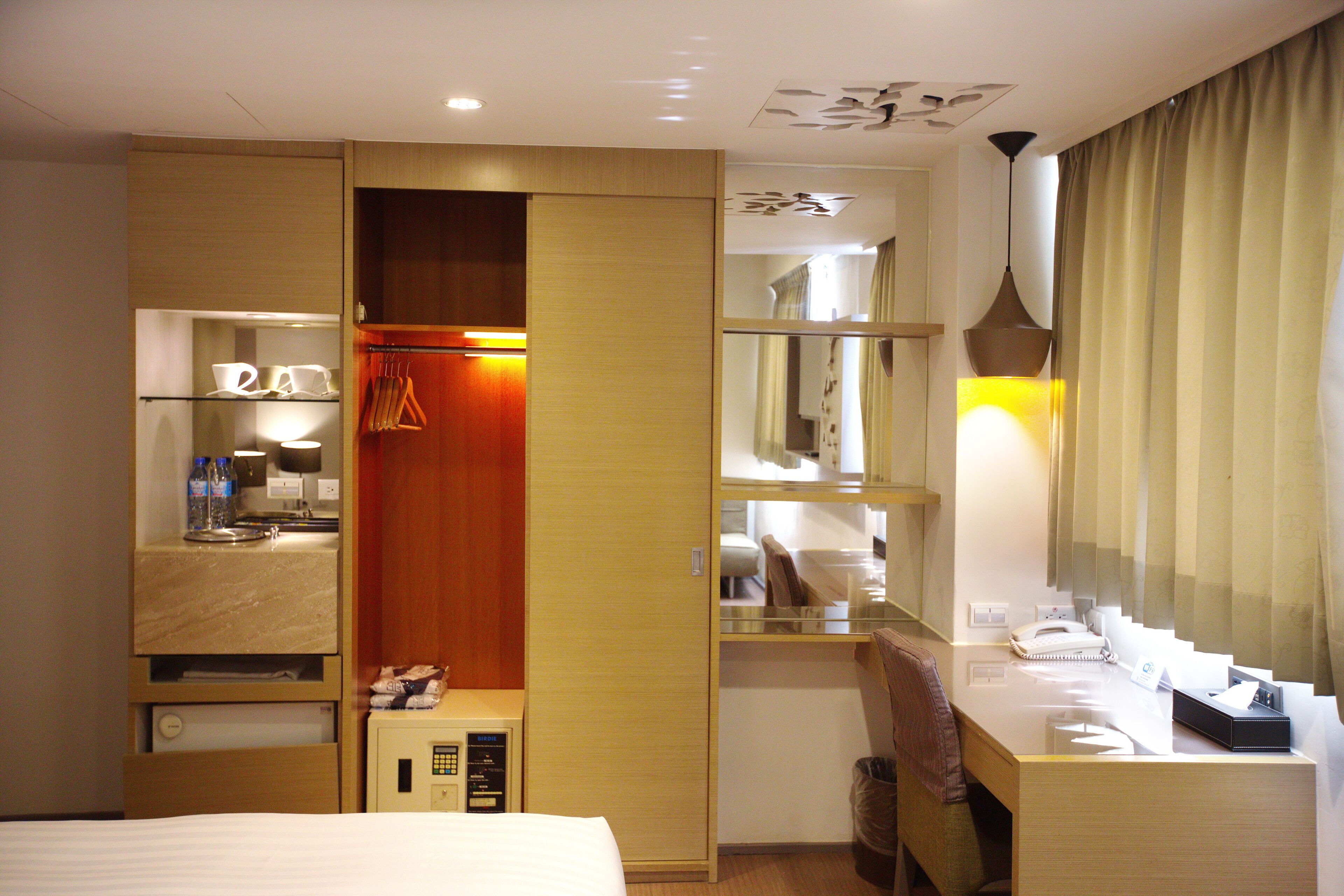 A hotel room with a bed, a desk with a lamp, a wardrobe, and a mirror.