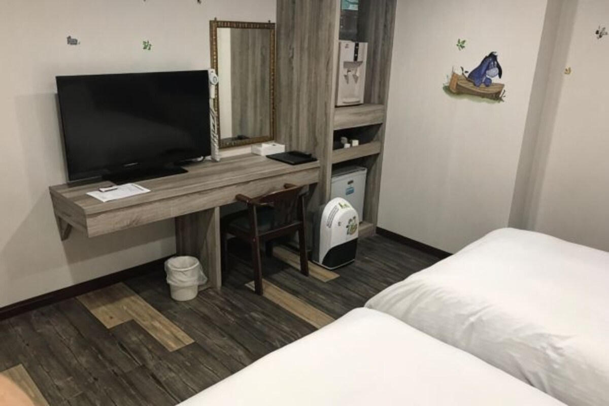 A hotel room with a bed, a desk with a computer, a chair, a television, and a small white bucket.