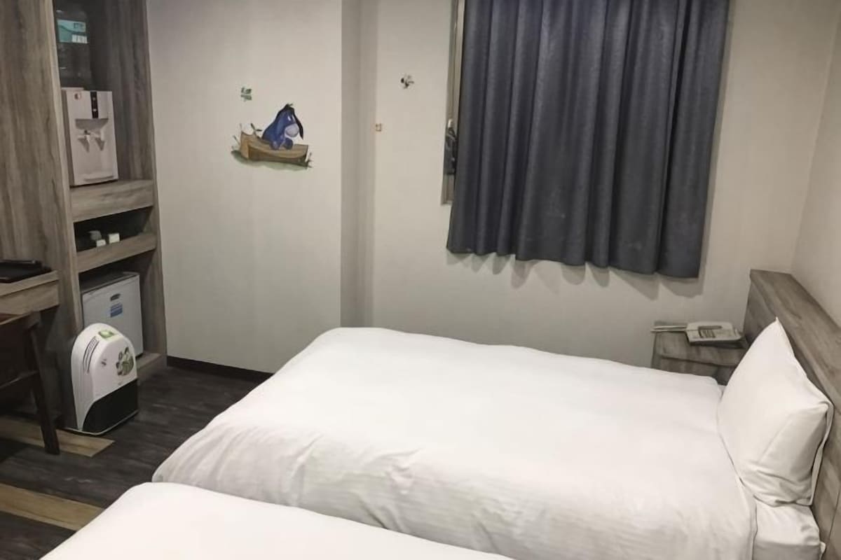 A hotel room with a bed, a desk with a computer, a small shelf with a bag, and a window with curtains.
