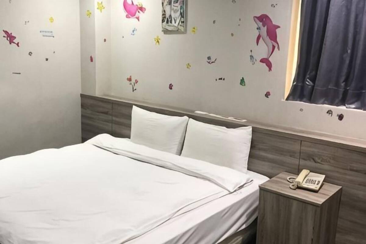 A bed with white bedding, a wooden bedside table with a phone, and a wall decorated with colorful stickers.