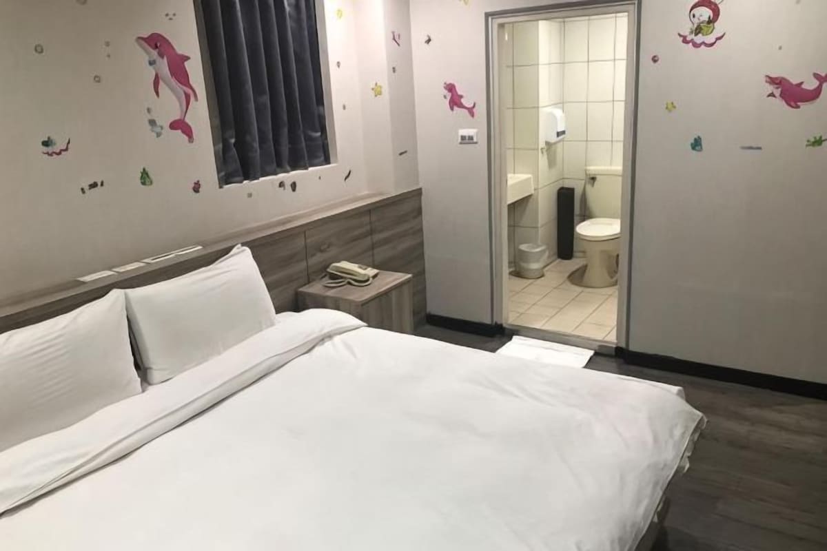 A hotel room with a bed, a nightstand, a window, and a view of the bathroom.