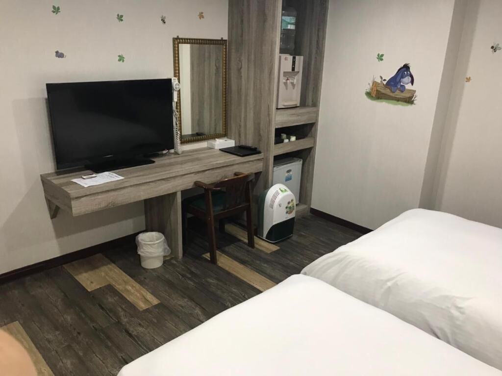 A hotel room with a bed, a desk with a TV, a mirror, a mini-fridge, and a trash bin.