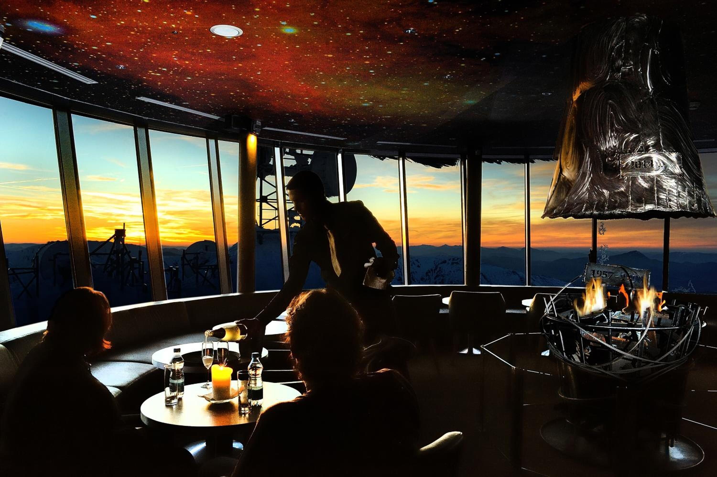 A restaurant with a view of the sunset, large windows, and a unique ceiling design.