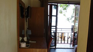 A compact hotel room with a wooden desk, a mirror, a small table, and a chair. A sliding door leads to an outdoor balcony with a view of the outdoors. - Phi Phi Green House (Ko Phi Phi)