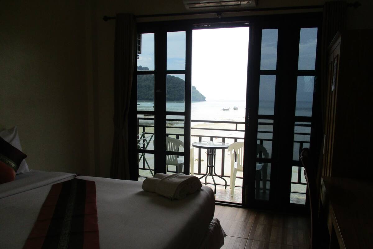 A hotel room with a bed, a view of the sea through large glass doors, and a balcony with a table and chairs.