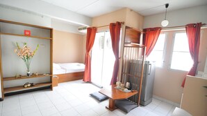 Quarto de hotel com cama, uma prateleira de madeira com objetos decorativos, uma mesinha com chaleira e um frigorífico.