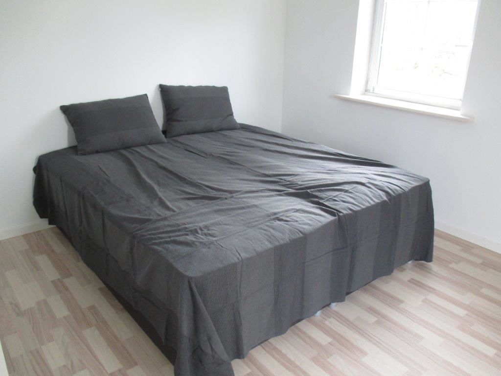 A bed with a dark bedspread and two pillows in a room with light wooden flooring and a window.