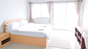 A bright hotel room with a large bed, a wooden nightstand, a desk, and a view through the glass door. - Greenville Serviced Apartment (Khlong Luang)