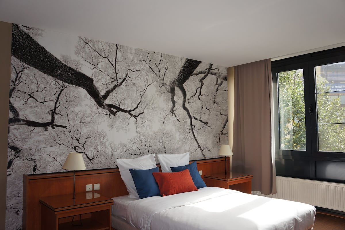 Un dormitorio con una cama, dos mesitas de noche, un ventanal grande y un mural en la pared.