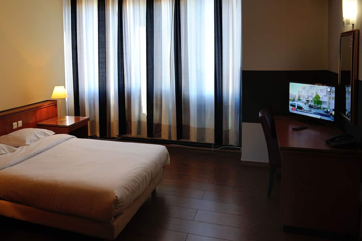Una habitación de hotel con una cama, dos lámparas de noche, un escritorio con un televisor y una ventana con cortinas.