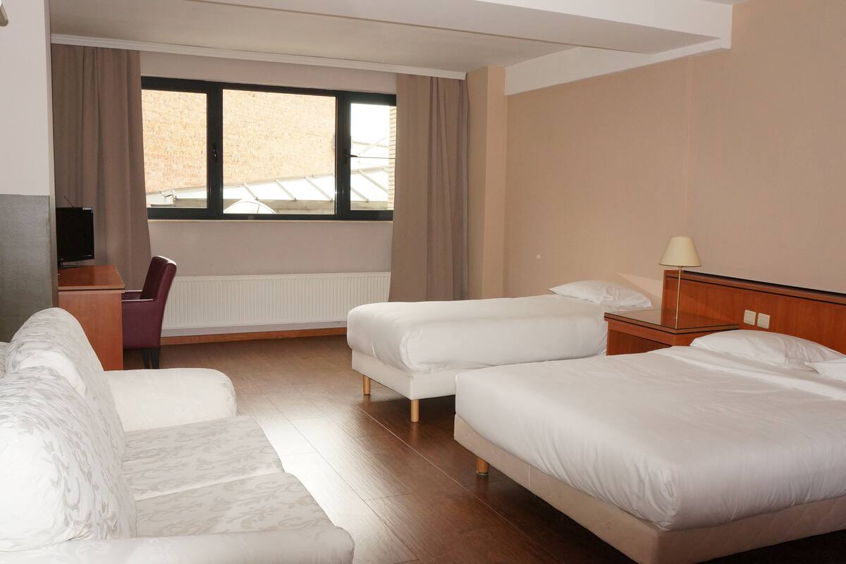 Habitación de hotel con sofá, dos camas individuales, escritorio con silla, lámpara y ventana con cortinas.