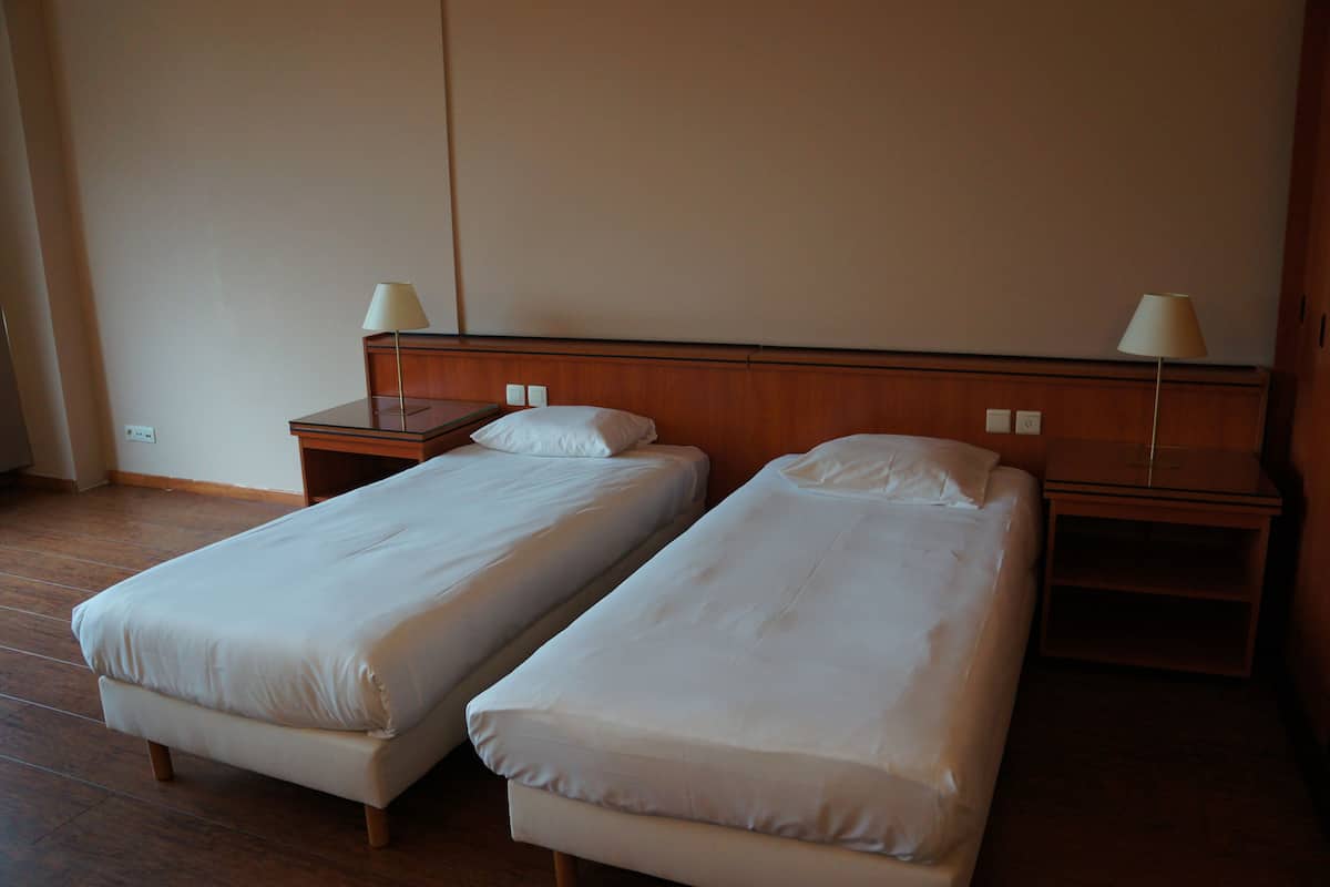 Habitación de hotel con dos camas individuales, cabeceros de madera, mesitas de noche con lámparas y una pared lisa.