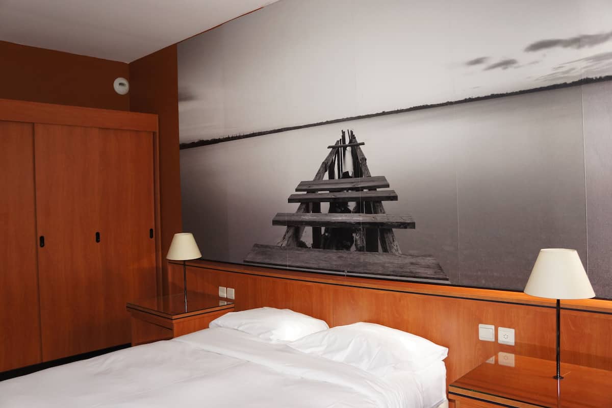 Habitación de hotel con una cama grande, cabecera de madera, dos mesitas de noche con lámparas y un mural grande con un muelle de madera y agua tranquila.