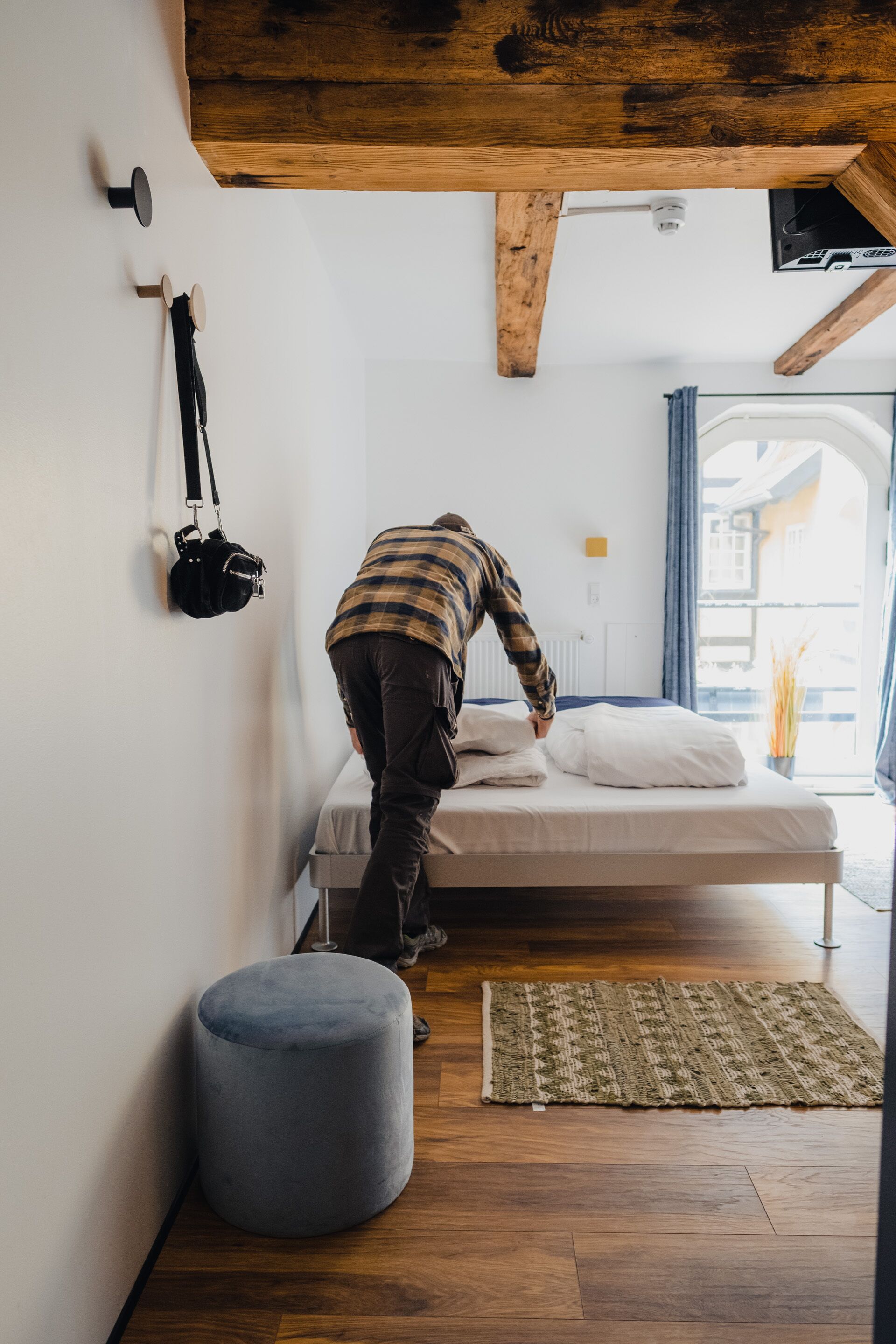 Un dormitorio con una cama, un reposapiés gris, techo de madera y una ventana con cortinas.