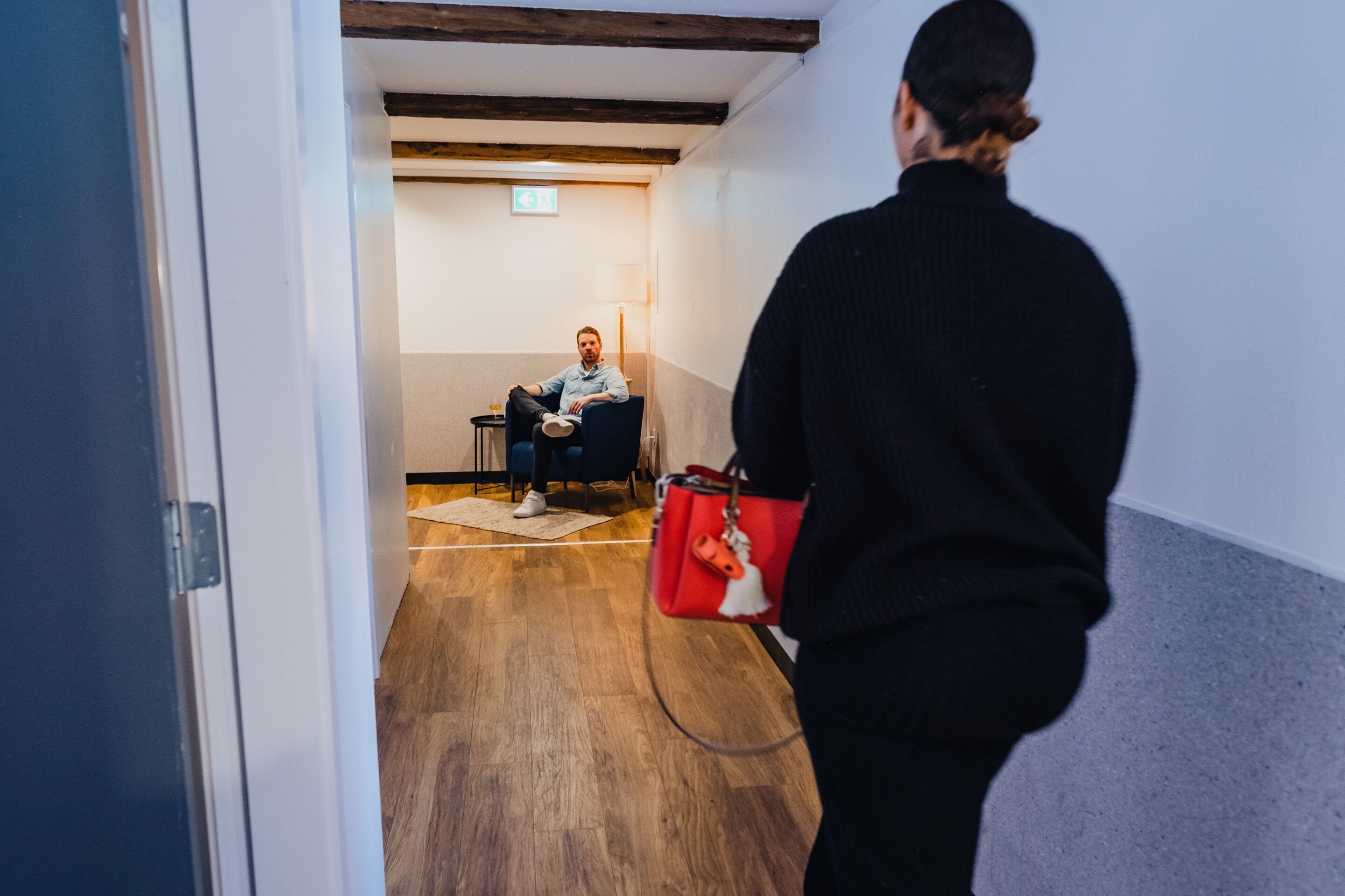 En person som går genom en korridor med trägolv, en person som sitter på en stol och en röd väska.