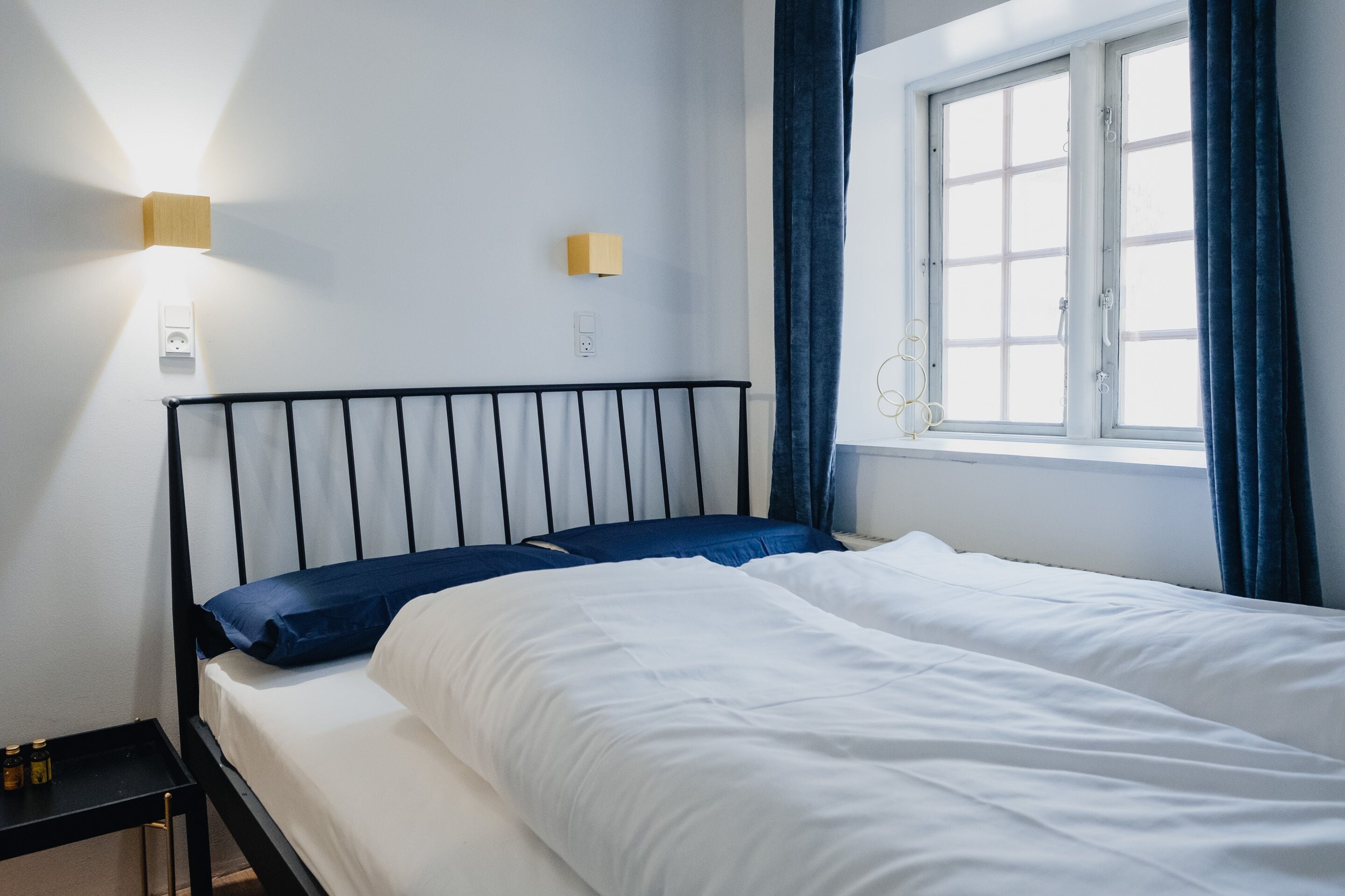 A neatly made bed with white and blue bedding, a black metal headboard, and two wall-mounted light fixtures.