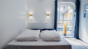 Ein ordentlich bezogenes Bett mit weißen Leinen in einem Zimmer mit einem weißen Heizkörper, zwei Wandleuchten und Blick auf ein Gebäude durch eine Tür.