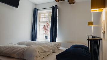 A bedroom with a bed, a window with curtains, a potted plant, and a wall-mounted TV.
