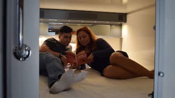 Una pareja recostada en una cama en un espacio pequeño y cerrado, con un área de kitchenette arriba.