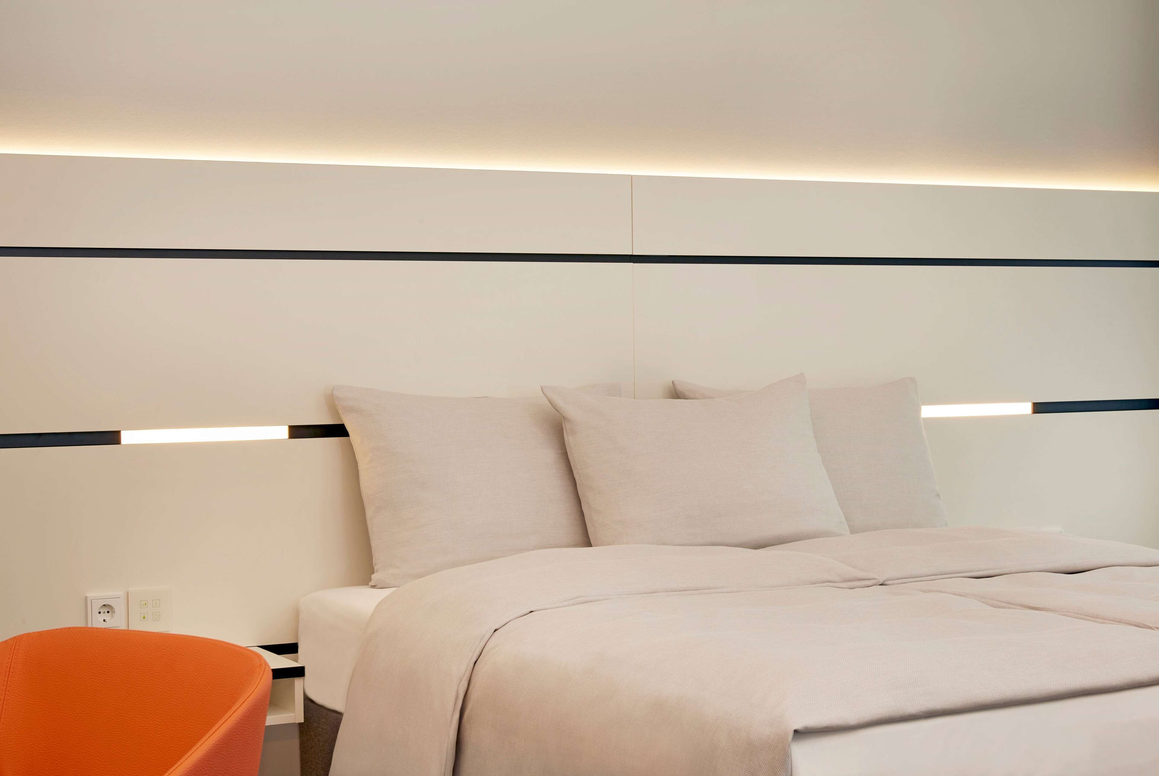 A modern hotel room with a neatly made bed, a small orange chair, and a wall-mounted light fixture.