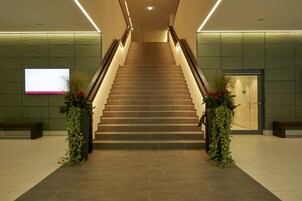Eine moderne Treppe, gesäumt von zwei Kübeln mit roten Blüten, führt zu einer Tür am Obergeschoss.