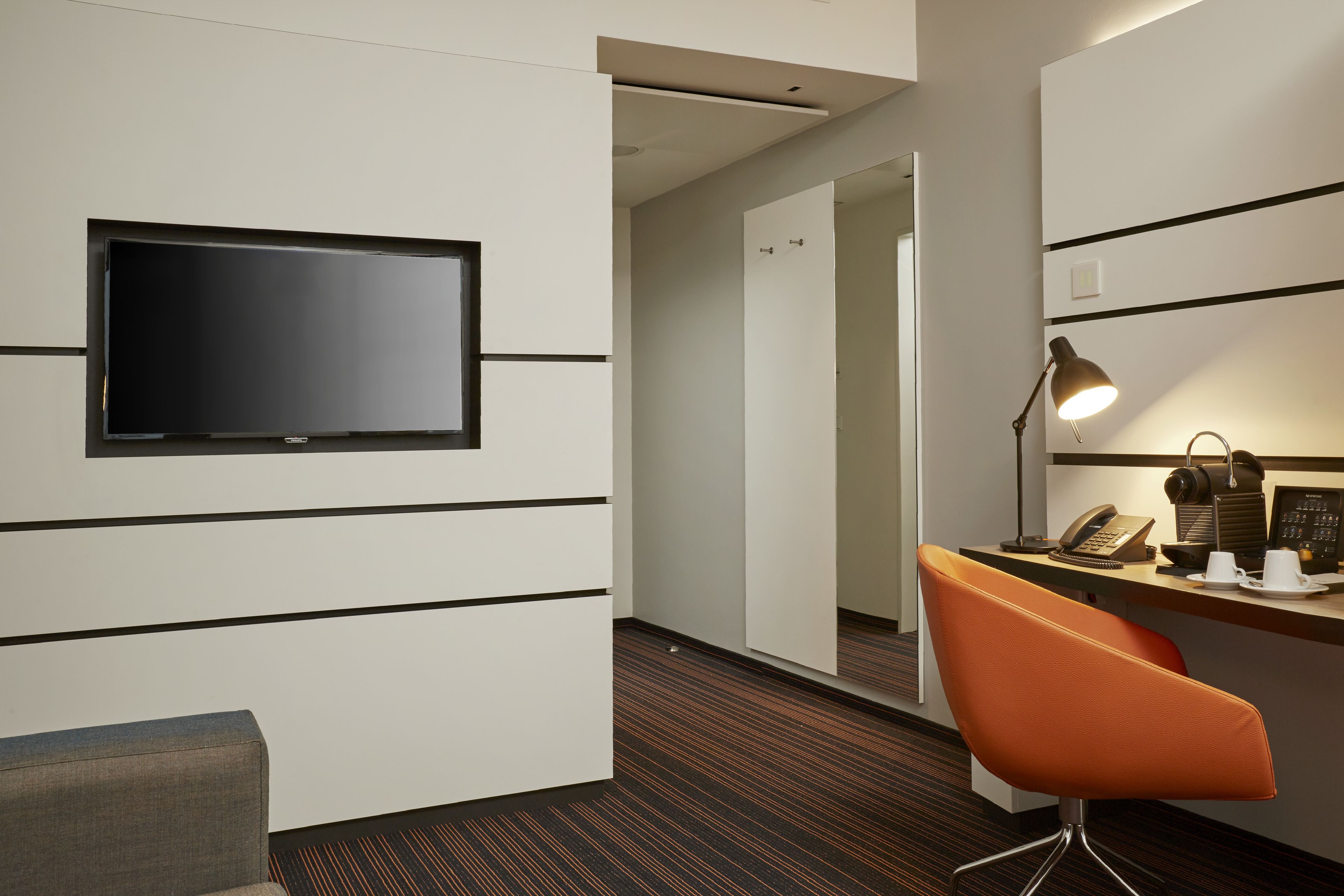 Una camera d'albergo con una TV a schermo piatto montata a parete, una scrivania con telefono e lampada, e una poltrona arancione.