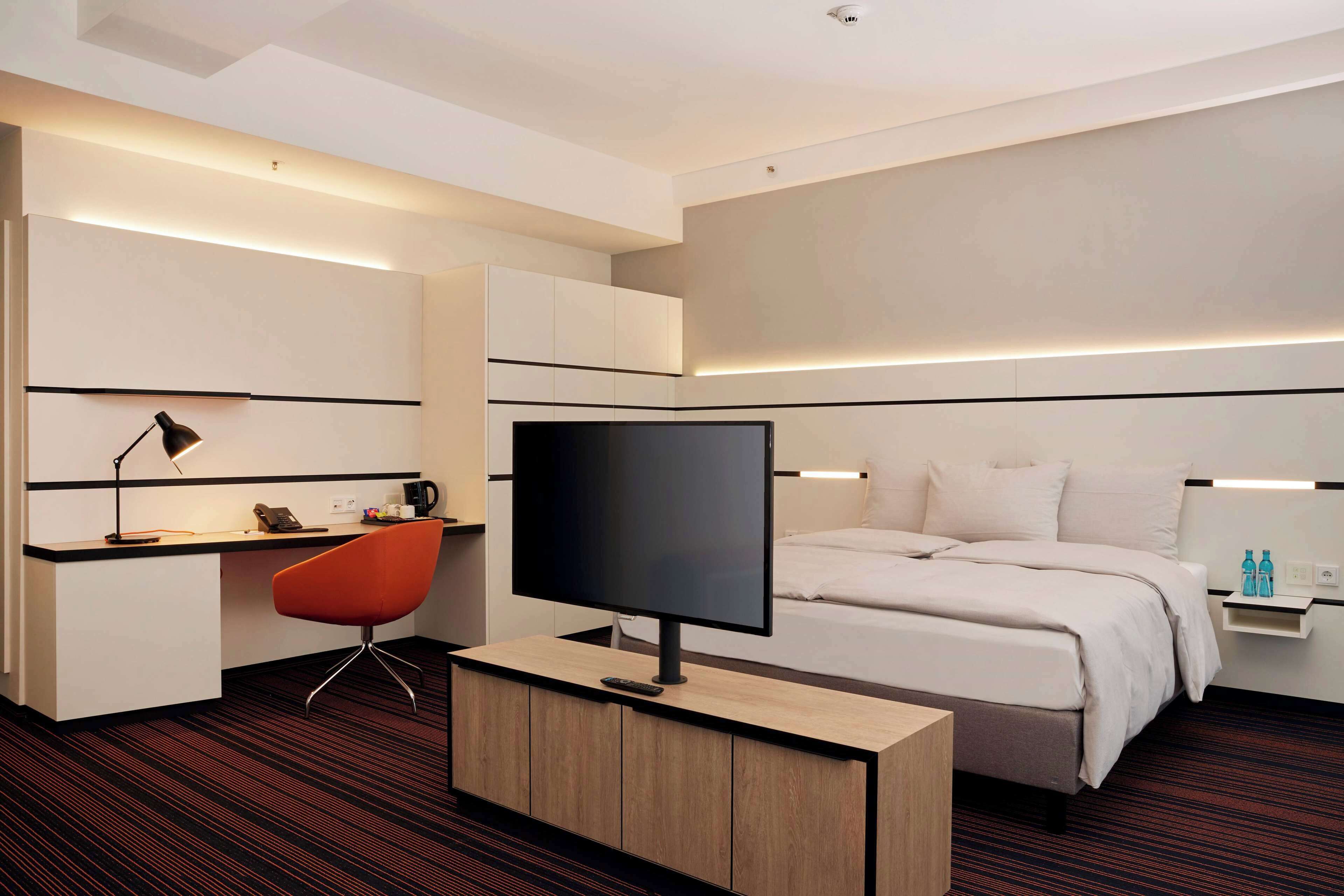 Una habitación de hotel moderna con una cama, un escritorio con computadora, un televisor y una silla.