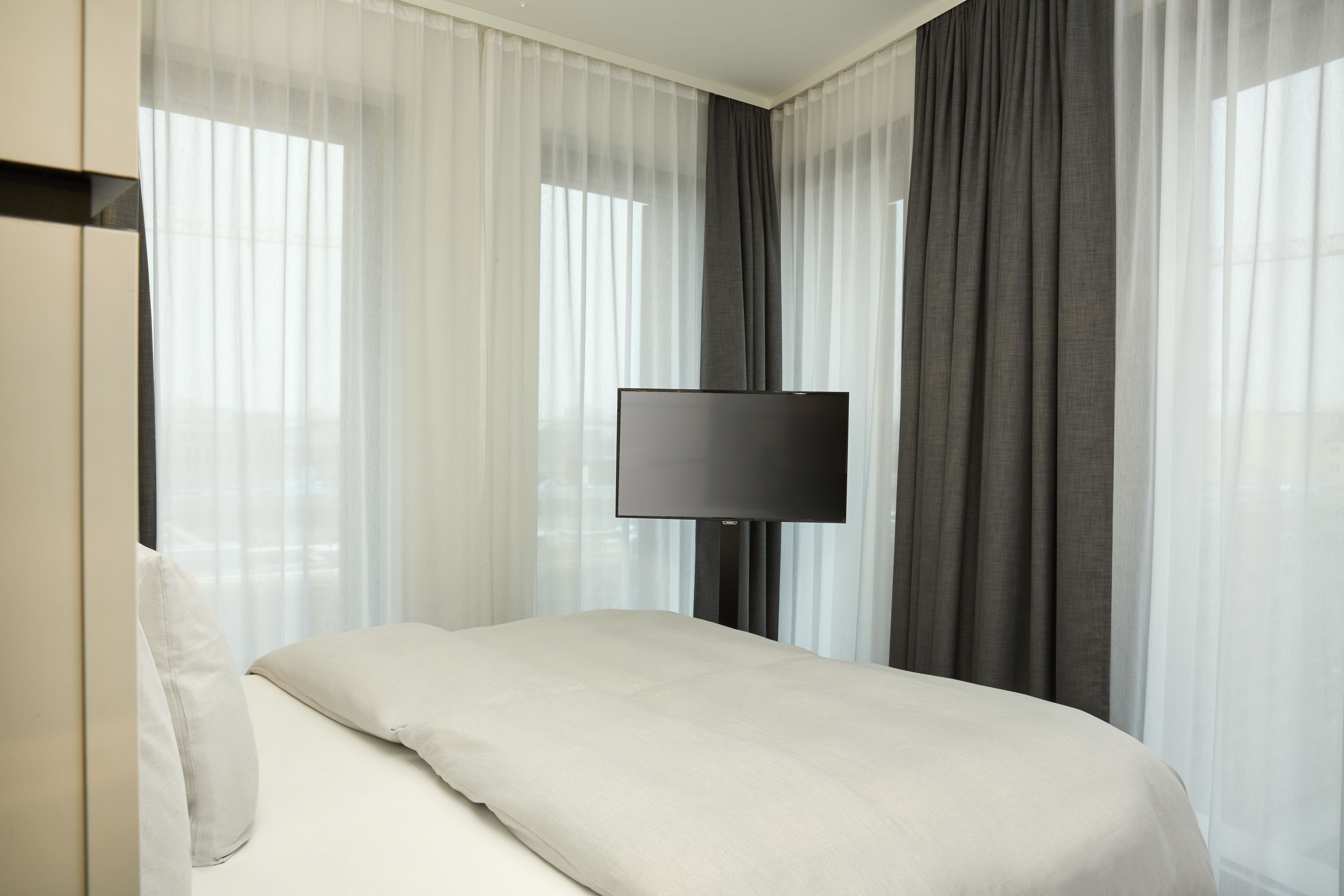 Una habitación de hotel con cama, televisión de pantalla plana, cortinas traslúcidas y una cortina oscura.
