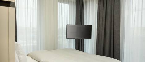 Una habitación de hotel con cama, televisión de pantalla plana, cortinas traslúcidas y una cortina oscura.