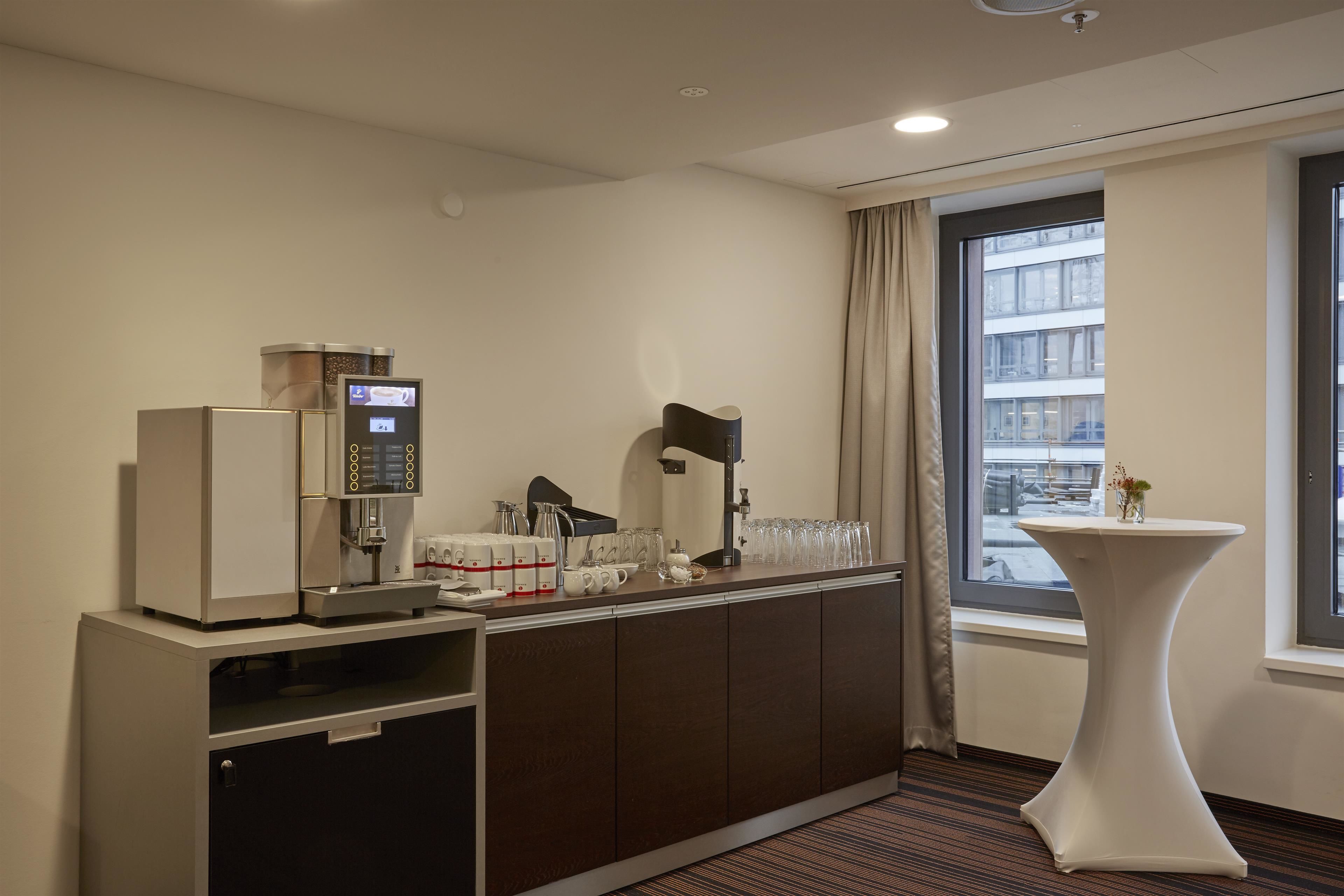 Une chambre d’hôtel équipée d’une kitchenette, d’une machine à café, d’une petite table avec un vase et d’une fenêtre donnant sur des immeubles.